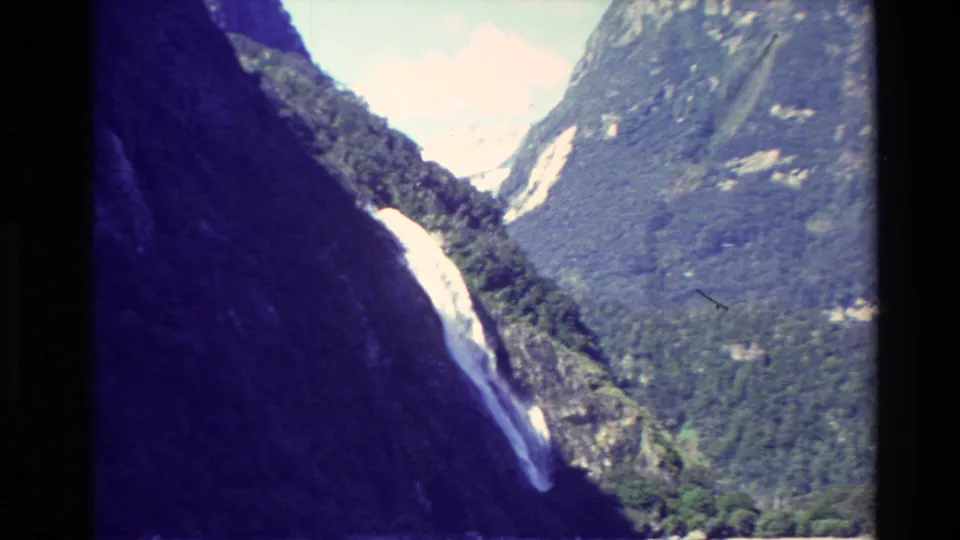 1982 New Zealand 8mm frame showing a waterfall descending through a narrow mountain ravine with forested slopes and bright sky.