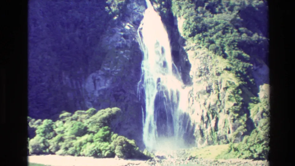 1982 New Zealand 8mm frame of a tall white waterfall falling over a rocky cliff with dense green forest at the base and side.