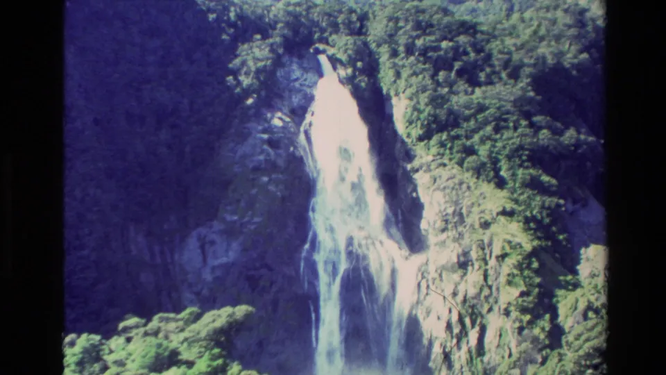 1982 New Zealand 8mm frame with a closer view of the waterfall spilling over dark rock, surrounded by green vegetation and cliff textures.