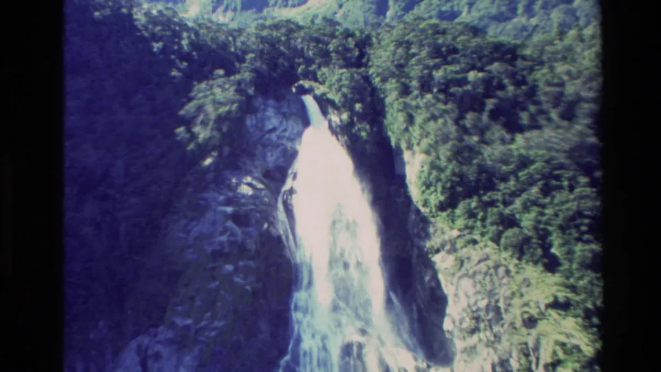 1982 New Zealand 8mm frame showing the waterfall framed by steep rocky slopes, dense forest, and bright daylight exposure.