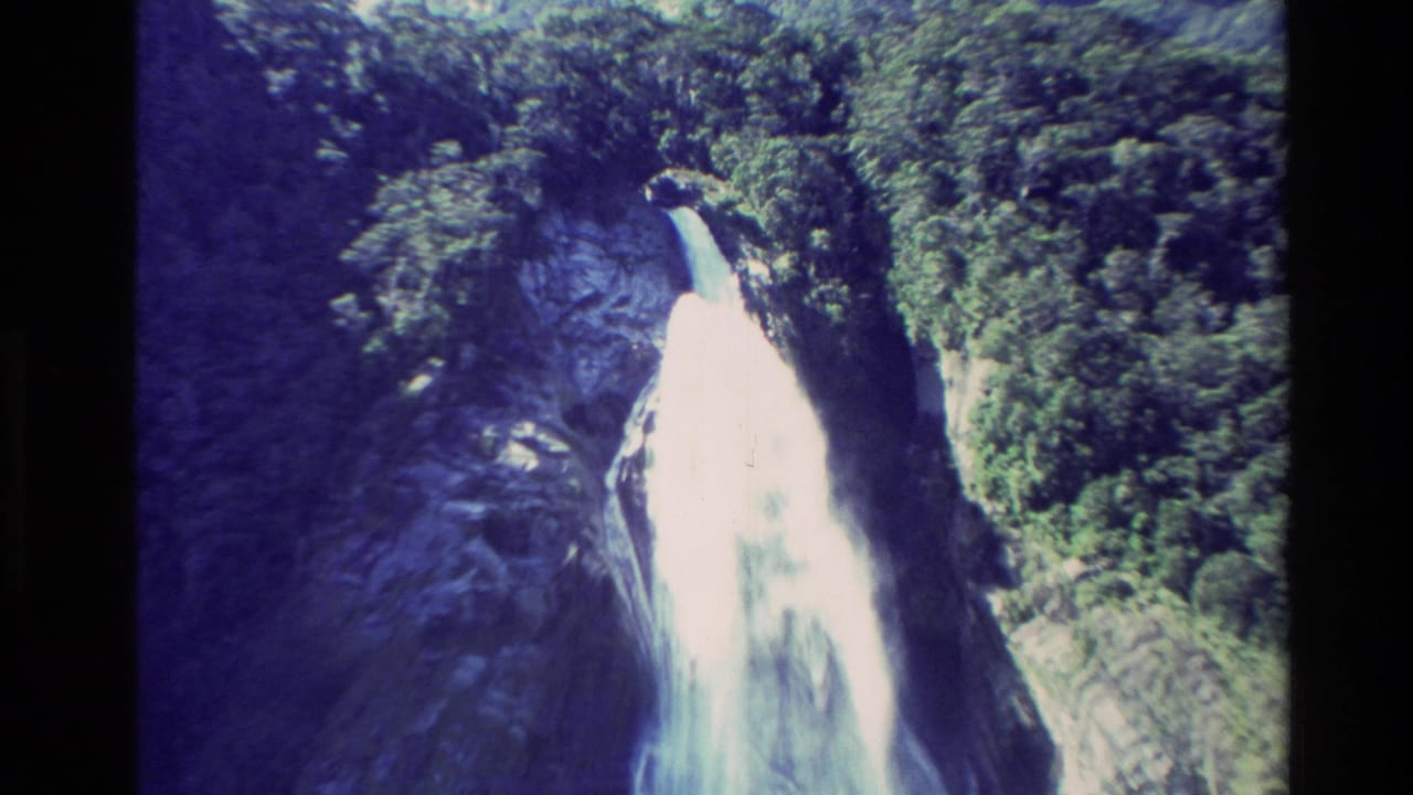 1982 New Zealand 8mm frame showing a bright waterfall descending through dense green forest and dark rock.