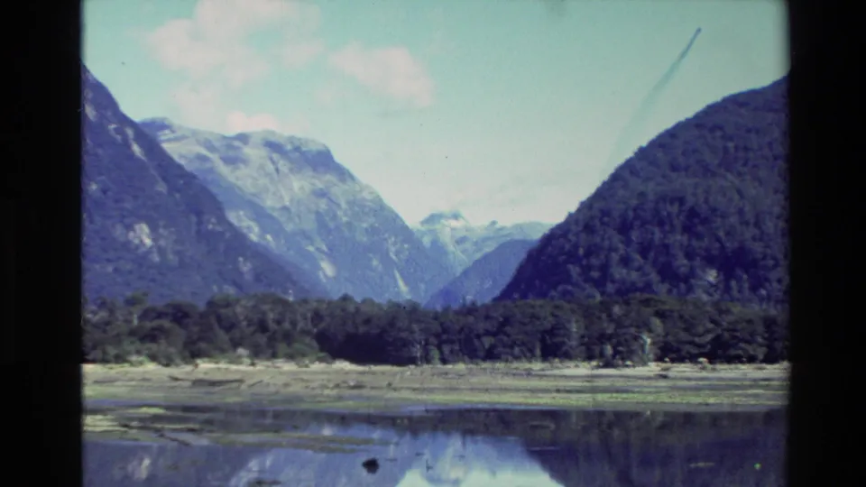 1982 New Zealand 8mm frame showing a calm lake or fjord below steep mountains and a partly cloudy sky.