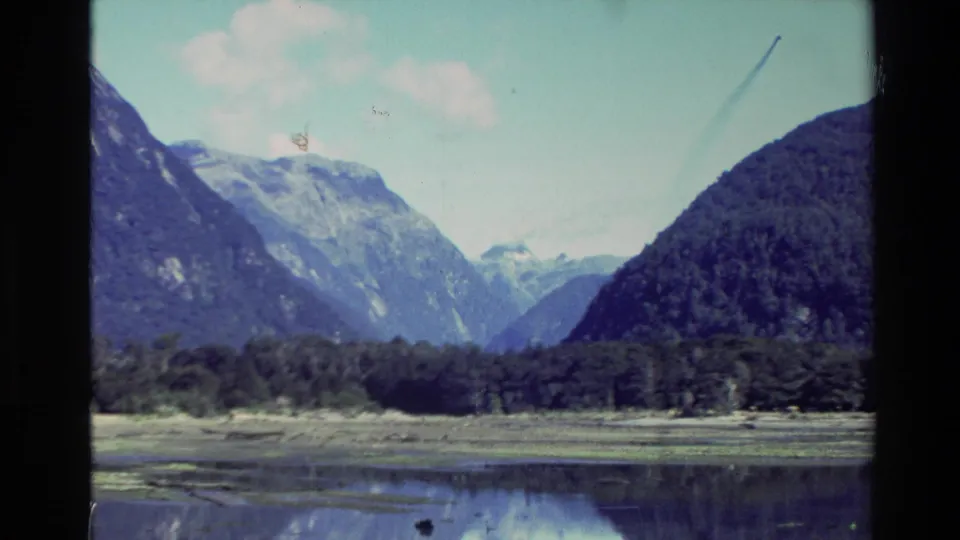 1982 New Zealand 8mm frame with a broad view of mountains, shoreline trees, and reflective water under pale sky.