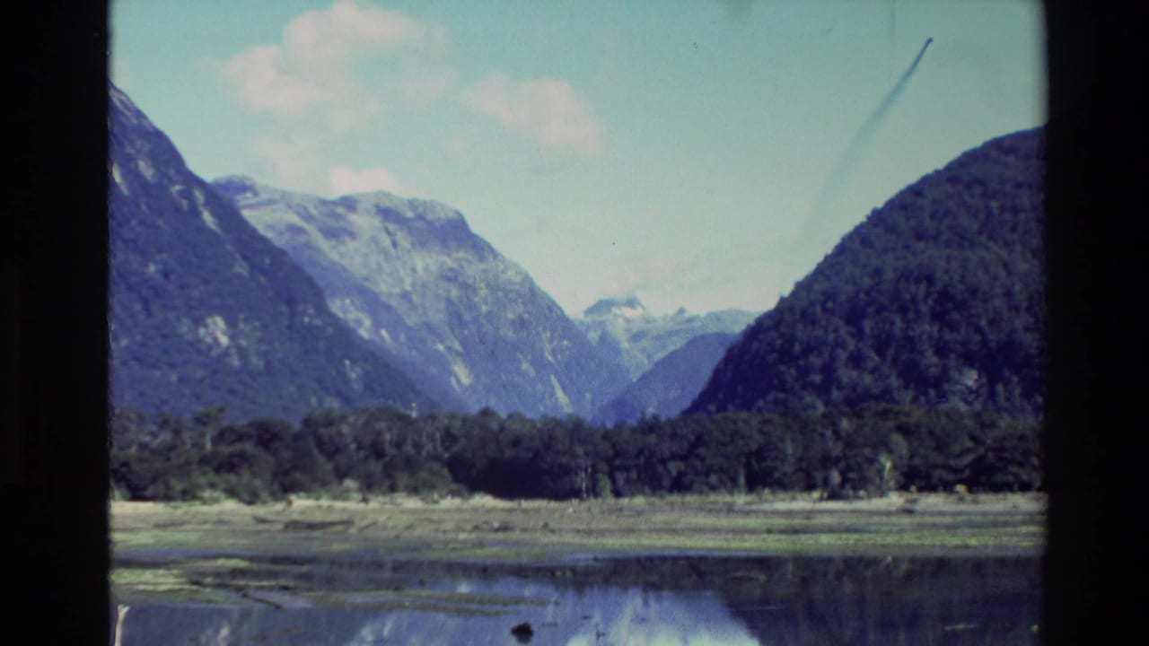 1982 New Zealand 8mm frame showing mountains, forested shoreline, and calm lake water under a bright sky.