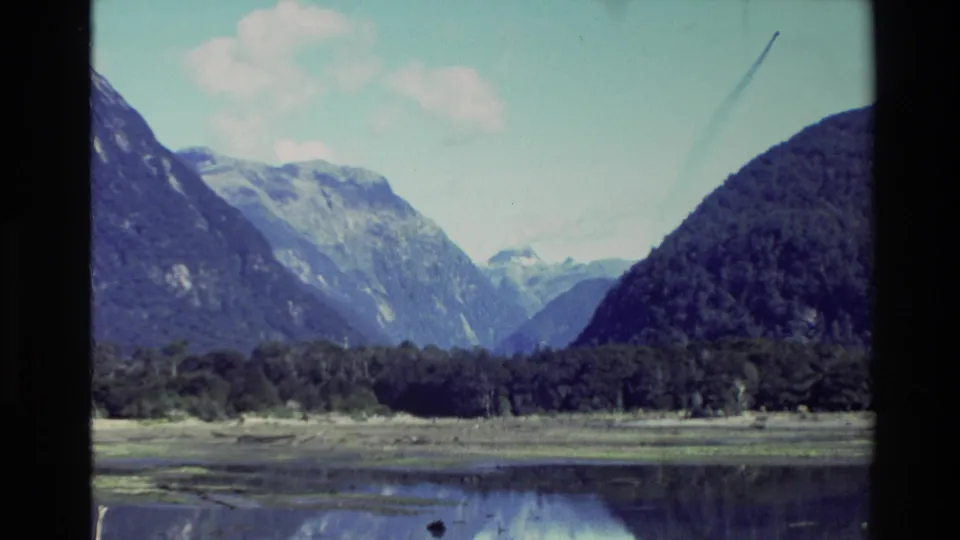 1982 New Zealand 8mm frame showing mountains, forested shoreline, and calm lake water under a bright sky.