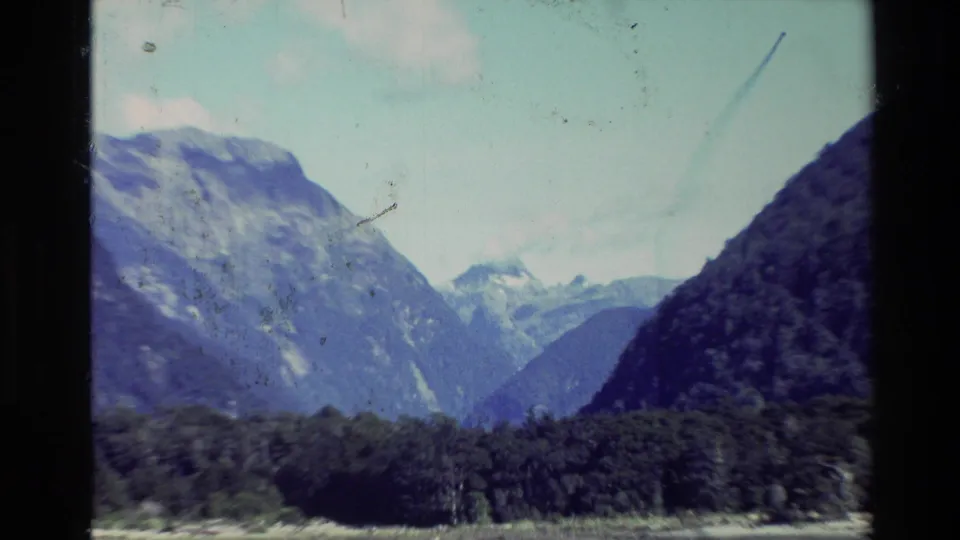 1982 New Zealand home-movie frame with steep mountain slopes, forested foreground, and distant snowy peaks.