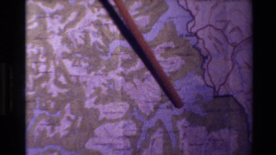 Close 8mm view of a wooden pointer tracing across a New Zealand topographical map with rivers or lake channels visible.