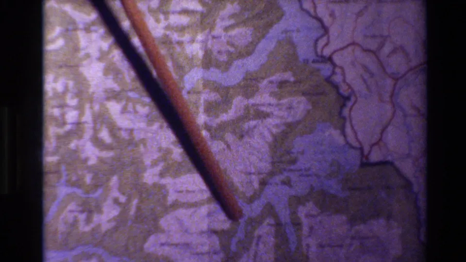 Projected 8mm frame of a wooden pointer laid diagonally across a topographical map, with water shapes and aged color visible.
