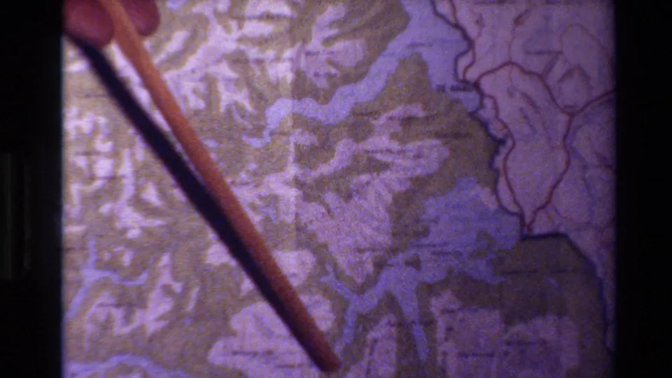 1982 New Zealand 8mm frame showing fingers guiding a wooden pointer across a topographical map with blue waterways.