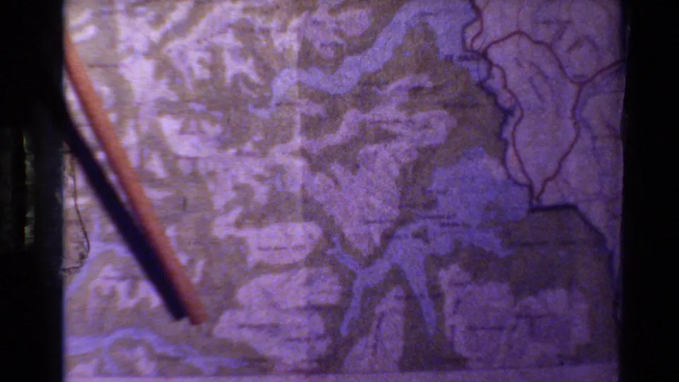 Home-movie frame of a stick angled across a topographic map, likely part of a travel route explanation in New Zealand.