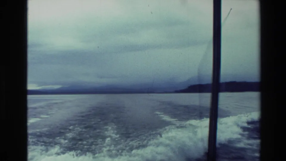 1982 New Zealand boat-ride view looking back over the wake toward distant shoreline and an overcast sky.