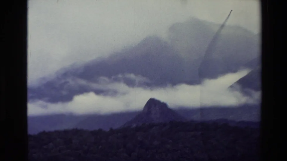 1982 New Zealand 8mm view from a moving boat, with a pointed dark hill, misty mountain ridges, and low clouds beyond the water.