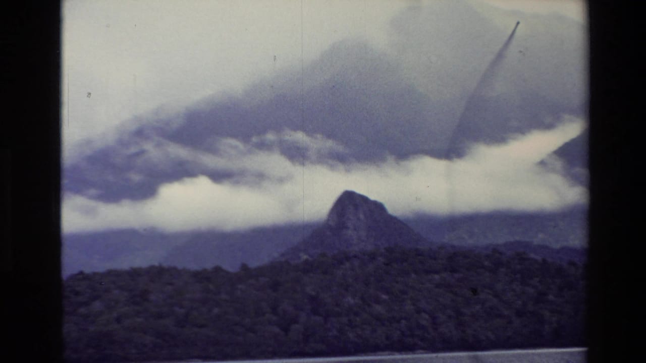 Dark mountain peak above forested slopes under low cloud, with a lake edge at the bottom.