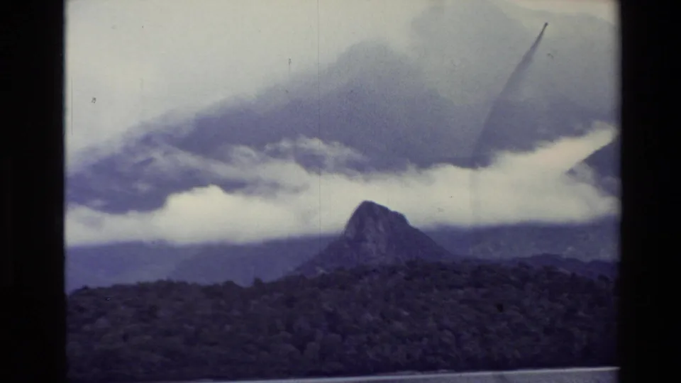 1982 New Zealand 8mm frame showing a dark mountain peak, forested slope, low clouds, and a narrow lake edge.