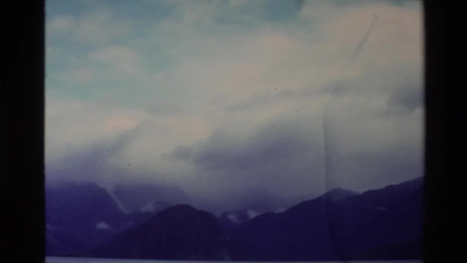 1982 New Zealand 8mm frame showing distant mountain silhouettes under heavy cloud, with muted color and visible film wear.