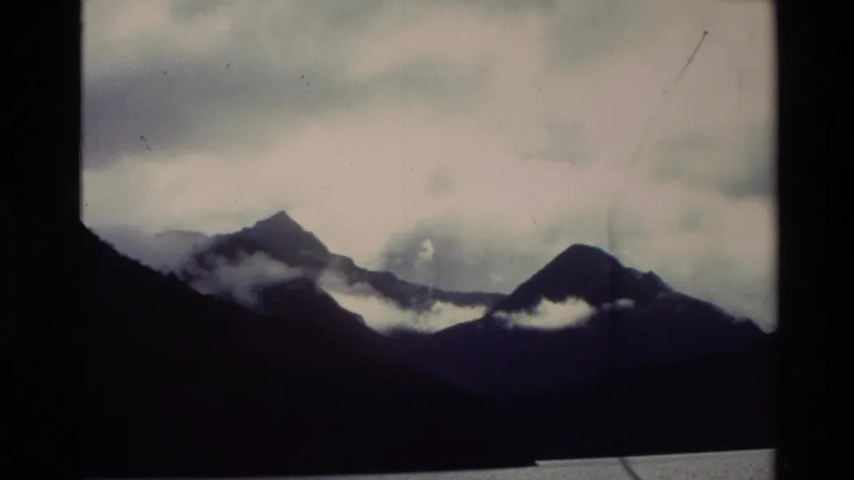 1982 New Zealand 8mm frame showing dark mountain forms, low cloud, a pale sky, and a thin lake edge.