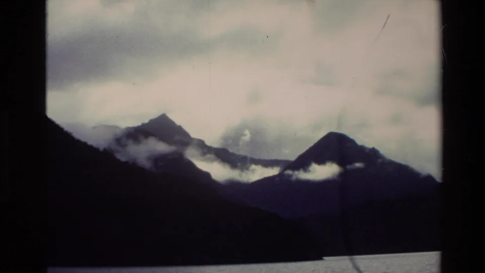 1982 New Zealand home-movie frame showing dark mountain silhouettes above calm water, with white mist drifting along the slopes.