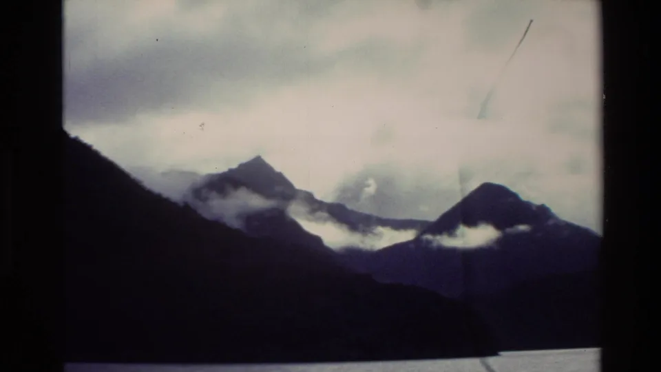 A Super 8 frame from New Zealand in 1982 shows mist crossing dark mountain slopes above a still lake or fjord.