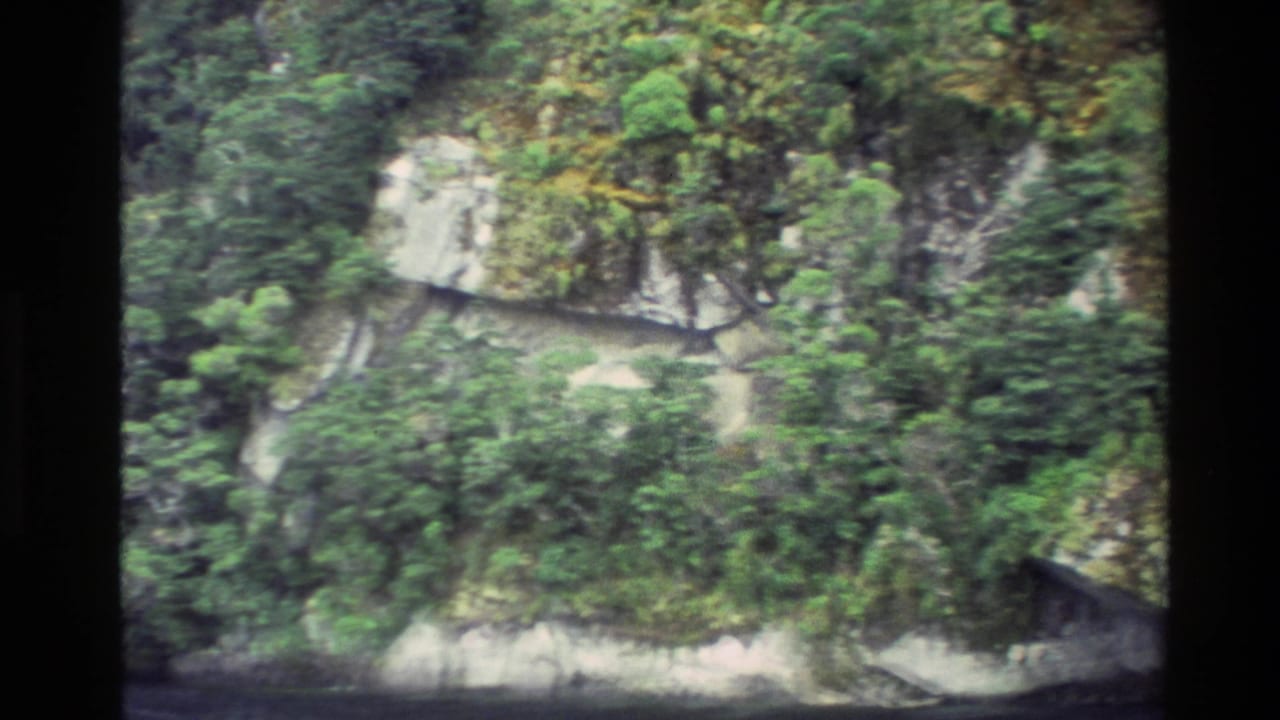 1982 New Zealand home-movie frame showing steep green vegetation and pale rock ledges above the water.