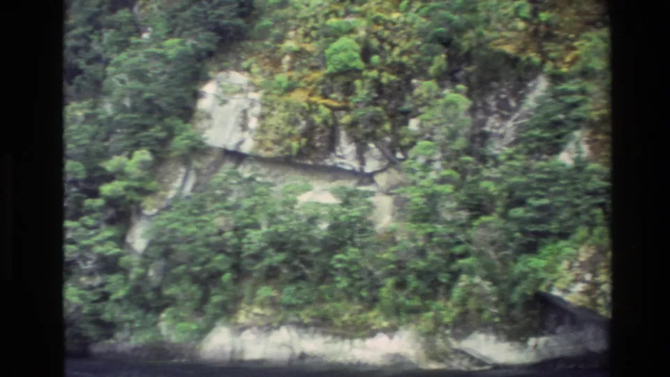 1982 New Zealand home-movie frame showing steep green vegetation and pale rock ledges above the water.