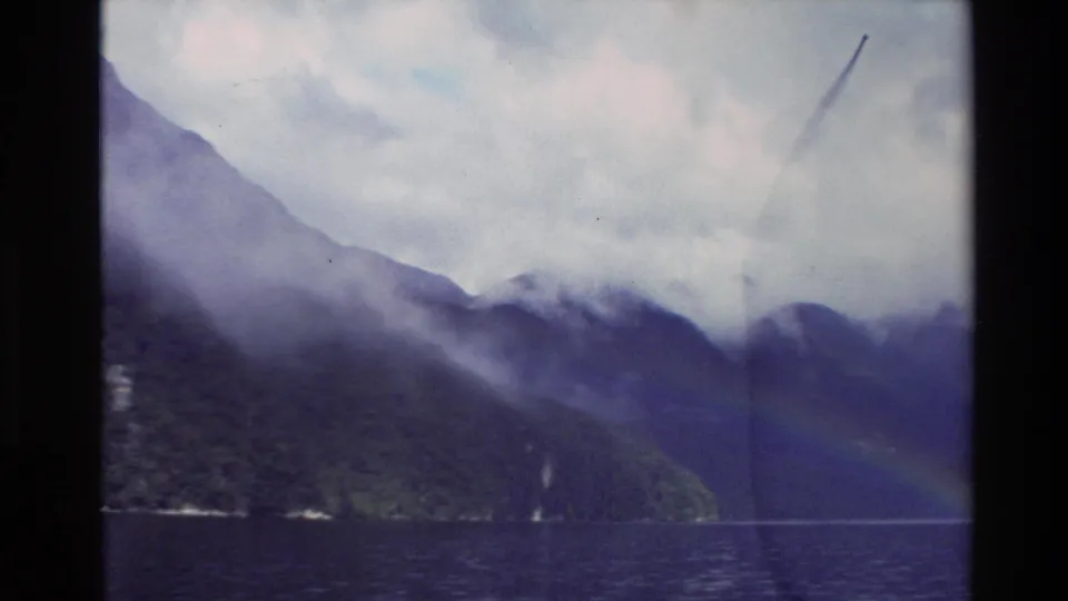 New Zealand 1982 8mm frame showing a misty fjord-like water view with mountain silhouettes, shoreline, and a faint rainbow.