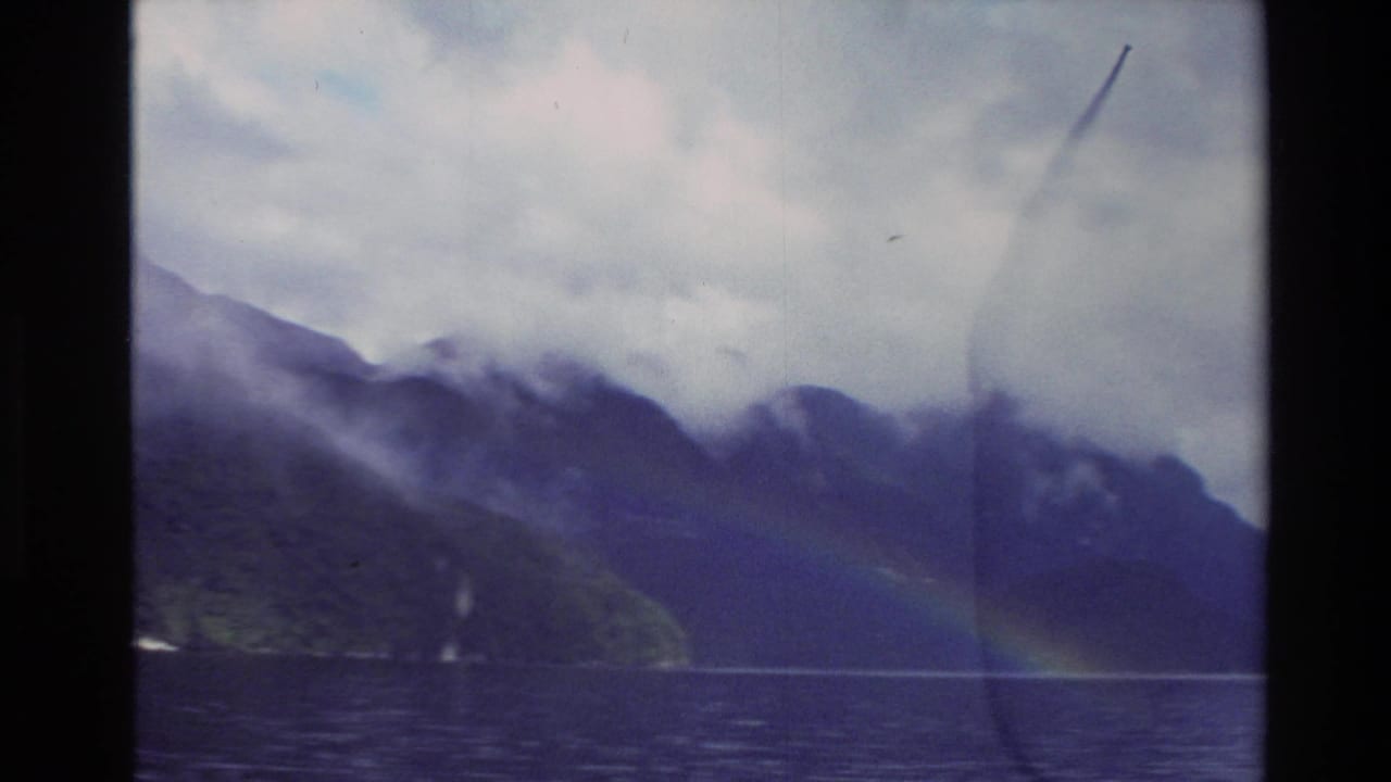 1982 New Zealand 8mm frame showing dark mountains, low cloud, lake water, and a faint rainbow near the far shore.