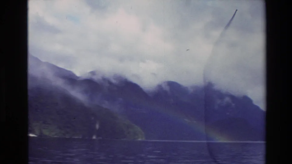 1982 New Zealand 8mm frame showing dark mountains, low cloud, lake water, and a faint rainbow near the far shore.