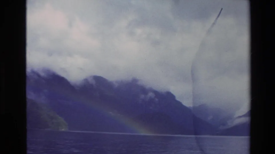 1982 New Zealand 8mm frame of a rainbow over a lake, with cloud-wrapped mountains and a dark film border.