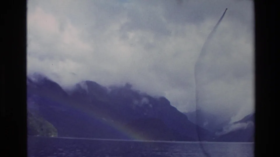 1982 New Zealand 8mm frame showing a lake below misted mountains, with a faint rainbow and aged film marks.