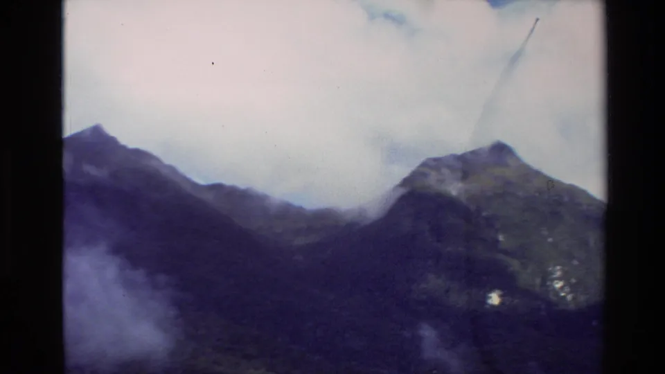 1982 New Zealand Super 8 frame with a steep mountain peak, cloud cover, and shadowed forested slopes.