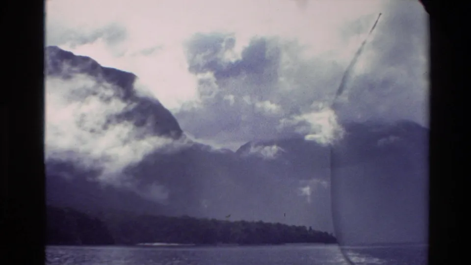 1982 New Zealand Super 8 frame showing low clouds over mountains, forested shoreline, and a strip of water.