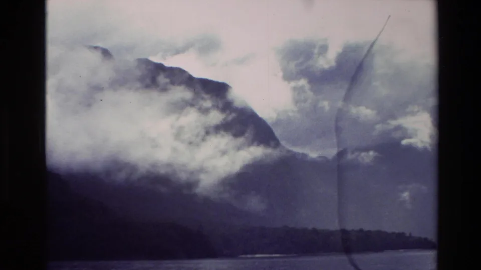 1982 New Zealand Super 8 frame with misty mountain slopes, a clouded valley, and water below.