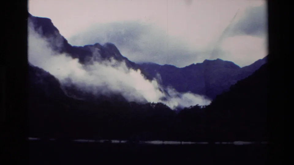 1982 New Zealand 8mm view of rugged dark mountains, low white mist, and a narrow strip of water under cloudy sky.