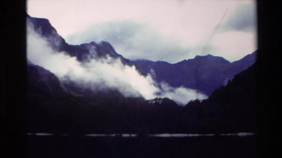 1982 New Zealand home-movie frame with mist pressed against steep mountain slopes and dark water in the foreground.