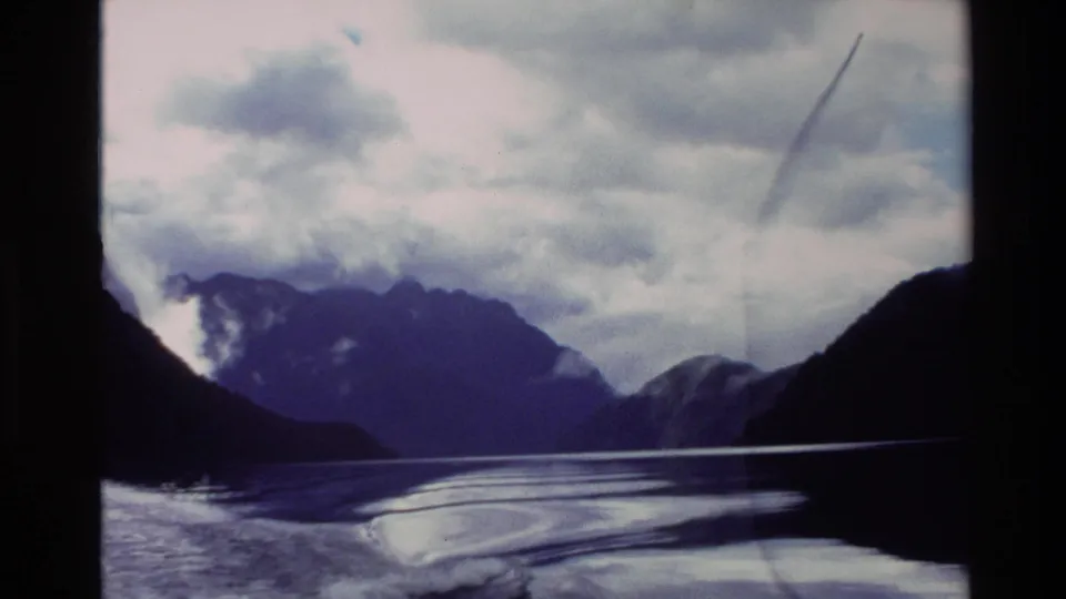 1982 New Zealand 8mm frame showing rippled water, distant mountain silhouettes, low cloud, and dark framing at the edges.
