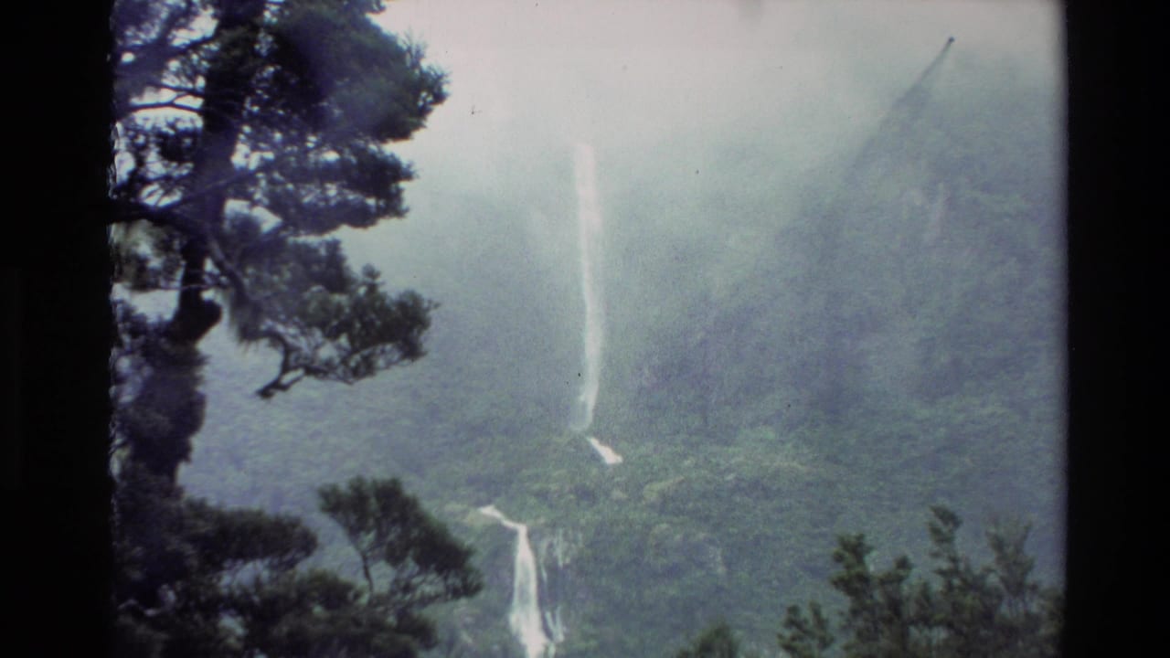 Misty green mountain forest with a tall white waterfall partly framed by dark tree branches.