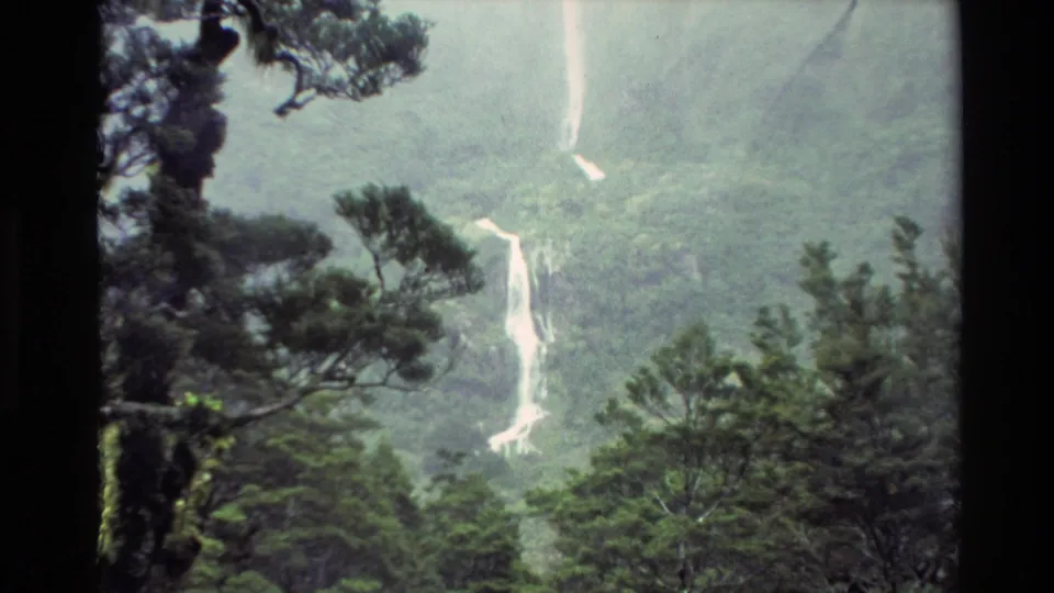 1982 New Zealand 8mm frame showing a waterfall and lower whitewater channel framed by dark branches and dense forest.