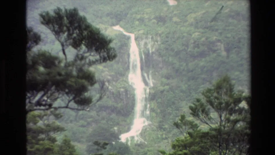 1982 New Zealand home-movie frame with a mist-softened waterfall, lower cascade, and evergreen trees crossing the foreground.