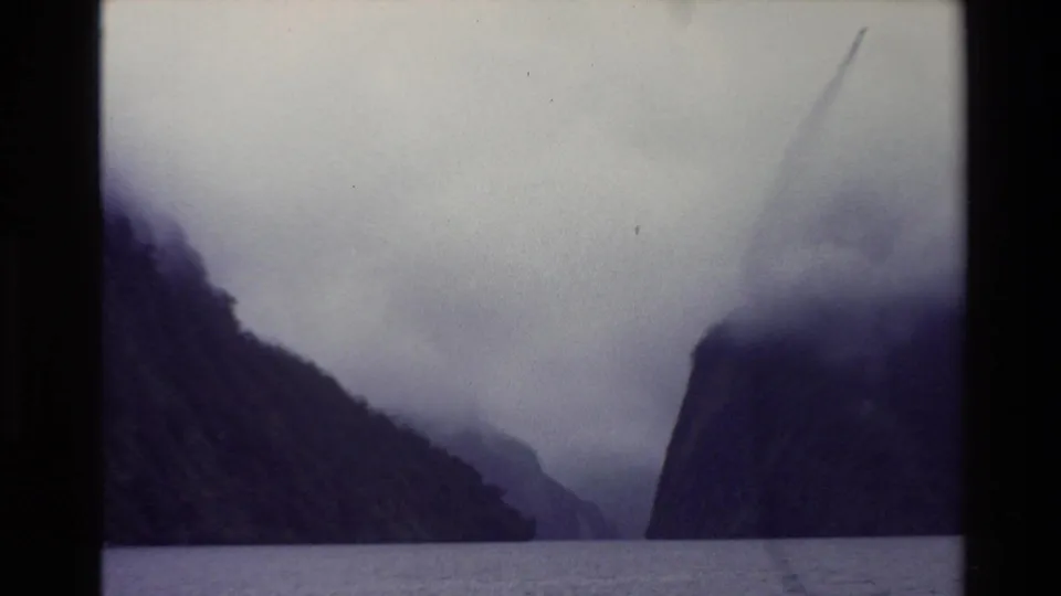 1982 New Zealand 8mm view of calm water between steep forested cliffs with low cloud covering the peaks.