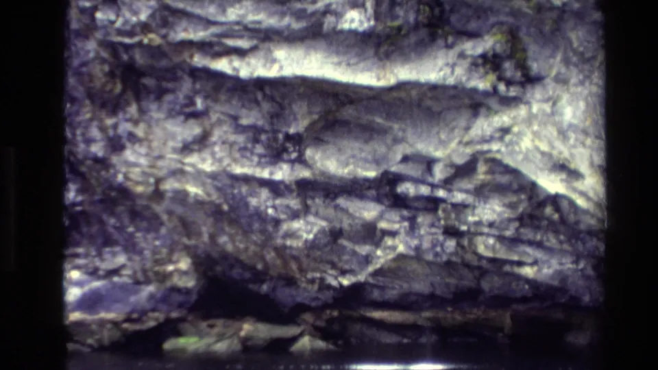 A closer 8mm frame of the rocky cliff edge and dark river water, with foliage visible near the upper edge.