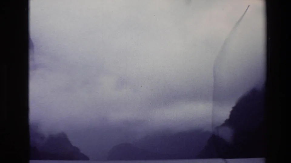 1982 New Zealand 8mm frame with distant mountain silhouettes, low cloud, water, and a vertical film mark near the right side.