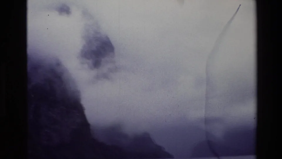 1982 New Zealand 8mm frame showing a dark mountain slope, low cloud, distant water, and soft exposure from the pan.