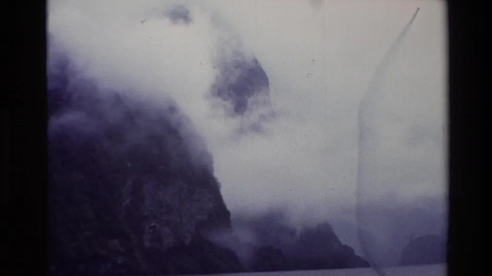 1982 New Zealand 8mm frame with a dark steep slope, drifting cloud, water, distant mountain forms, and a faint film-edge mark.