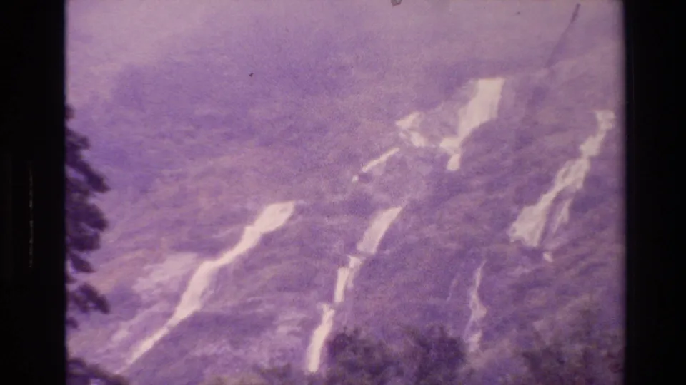 1982 New Zealand 8mm frame showing mountain slopes obscured by purple haze, with several pale smoke or plume trails crossing the terrain.