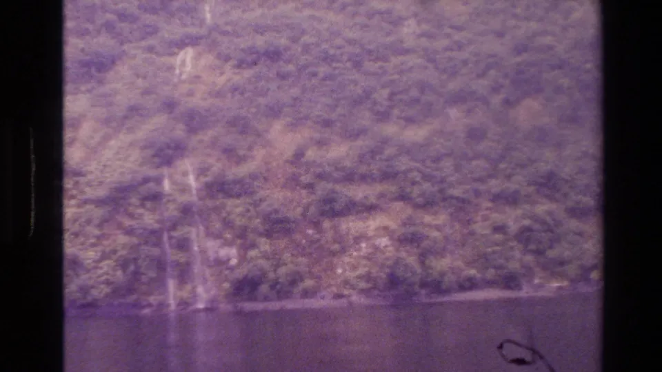 1982 New Zealand 8mm frame showing a distant forested slope, dark water or valley shadow, and a faint waterfall through purple film cast.