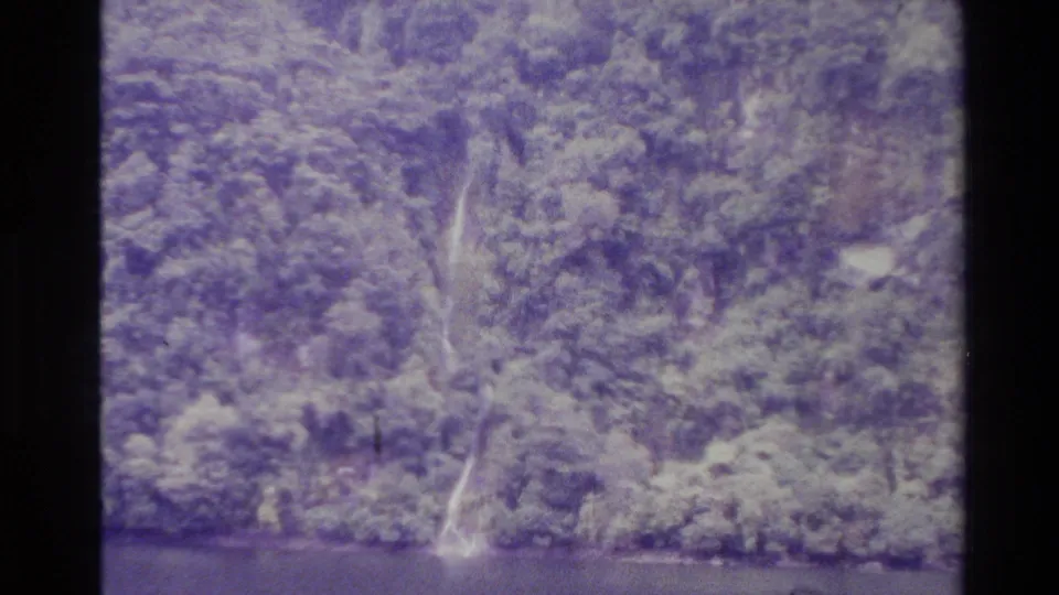 1982 New Zealand 8mm frame of a thin waterfall on a tree-covered hillside, seen from across calm water with soft film grain.