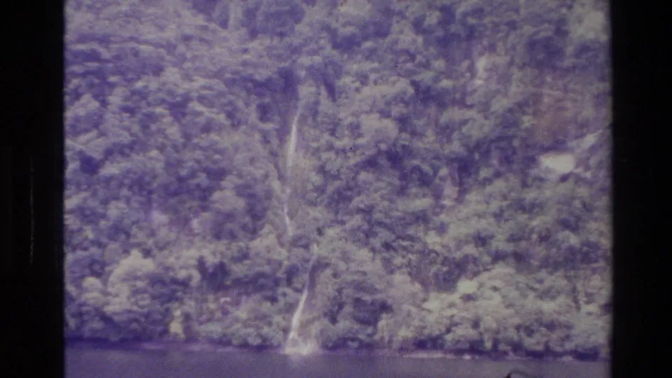 1982 New Zealand 8mm frame showing a waterfall descending through thick vegetation toward dark water at the base of the cliff.