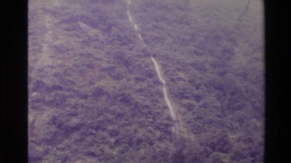 1982 New Zealand 8mm frame with a soft distant waterfall streaking down a forested cliff, heavily tinted by aged film color.