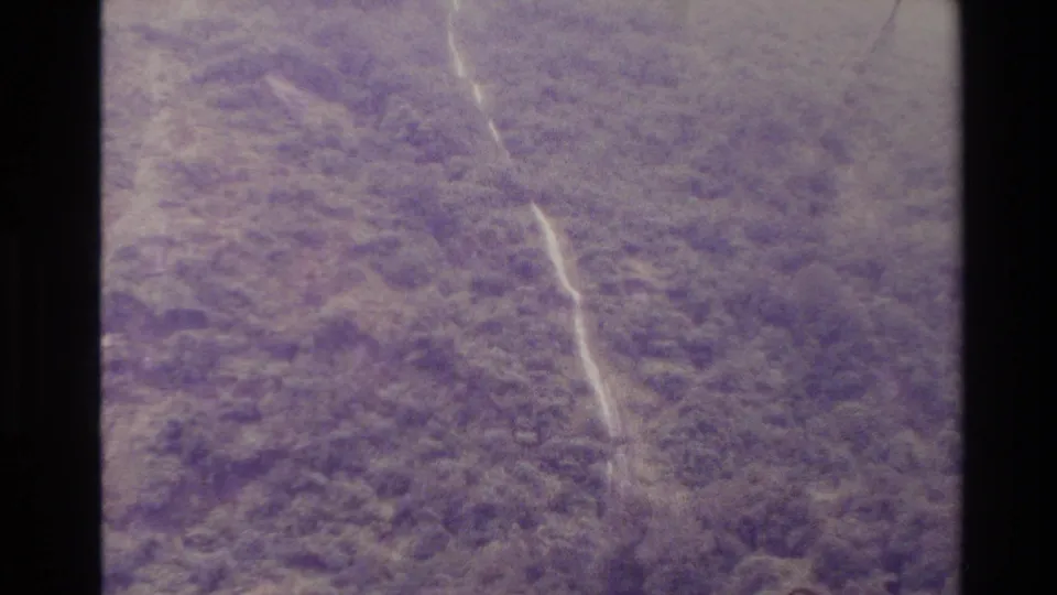 New Zealand 1982 aerial home-movie view of a wooded mountainside with a narrow pale watercourse and aged purple film color.