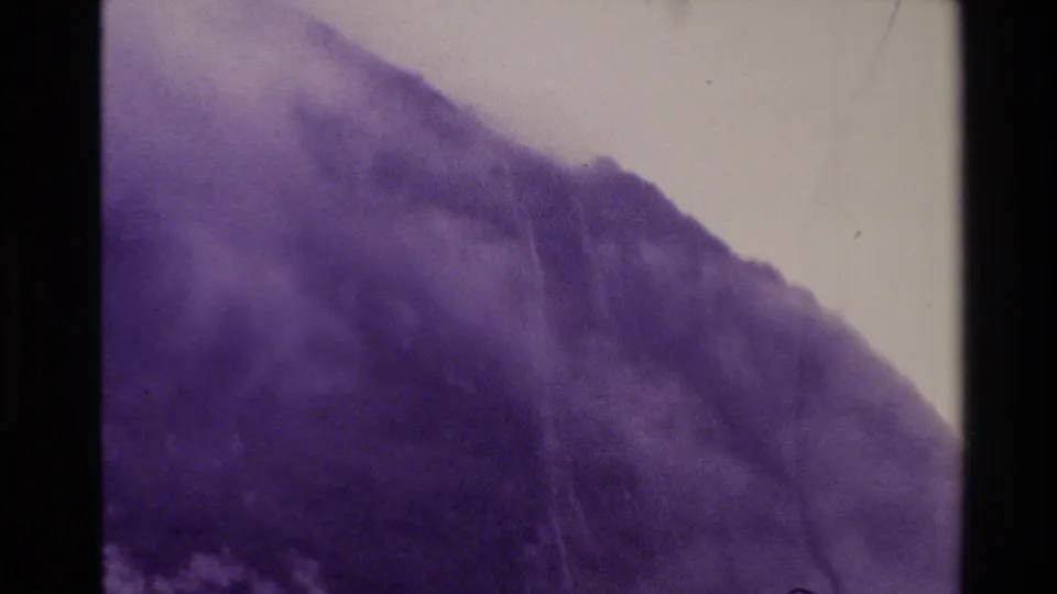 Aerial view of a mist-covered New Zealand mountain ridge in 1982 footage, softened by haze, grain, and purple color aging.